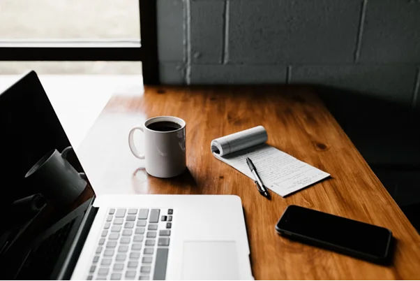 uma mesa de trabalho com um laptop aberto, uma caneca de café e um bloco de notas com anotações